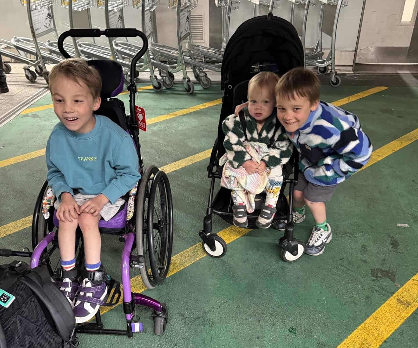 Frankie, Remy and Otis are side by side smiling widely in an airport. Frankie is sat in his purple wheelchair.