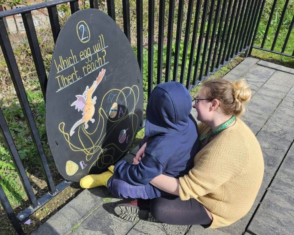 Matthew is sitting on his mother's lap, who is sat on the floor. They are looking at a big cut-out of a black egg, trying to complete the challenge written on it. The egg reads 'Which egg will Ilbert reach', with a picture of a dragon following several multicoloured lines.