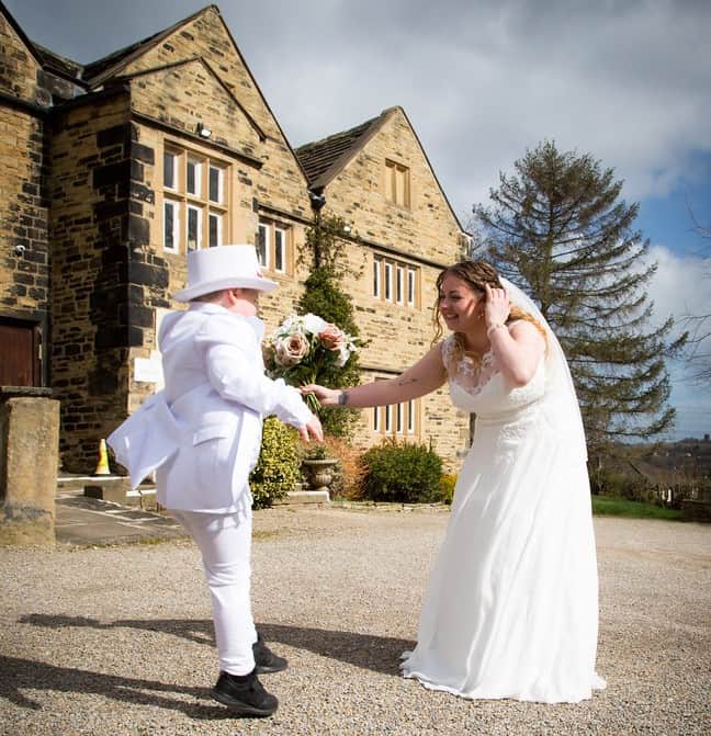 Matthew has his back to us, wearing a white suit and a white top hat. His mother, Lissi, is stood in front of him wearing a wedding dress and holding a bouquet of pink flowers.