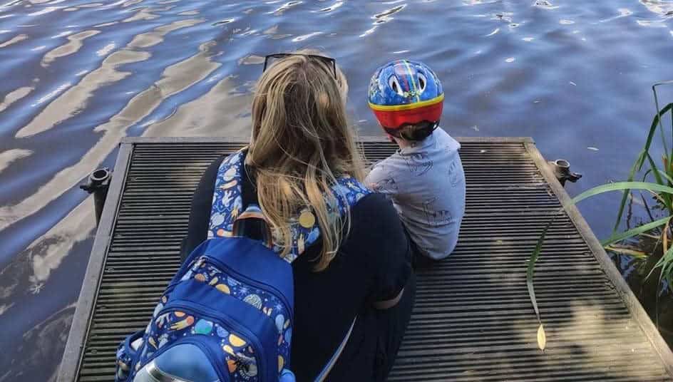 Lissi and Matthew are sitting on a wooden pier, on a lake, looking out at the water. They both have their back to us.
