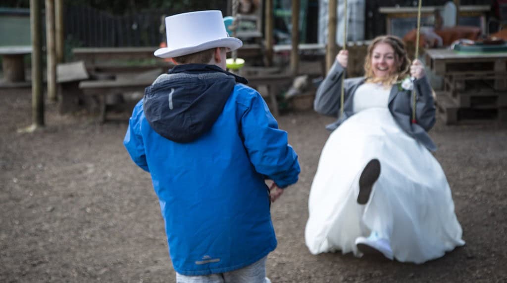 Matthew has his back to us, wearing a blue jacket and a white top hat. His mother Lissi is sat on a swing in front of him, wearing her wedding dress and smiling widely.