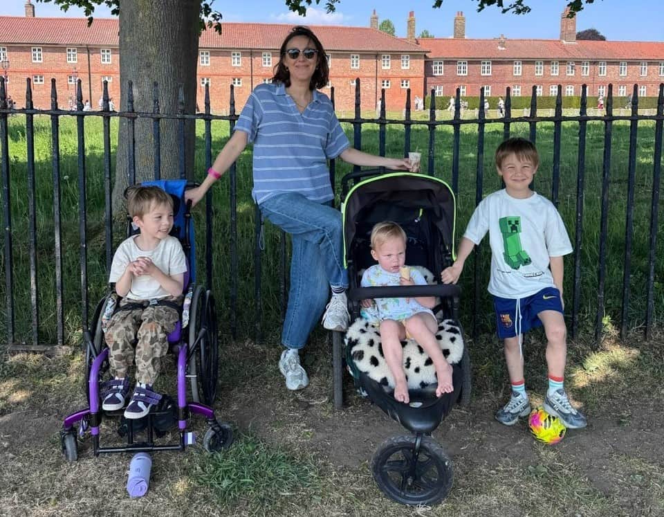 Melissa is smiling widely standing alongside her three boys. Frankie is to her left, in his purple wheelchair, and Otis is stood alongside Remy in his wheelchair, to Melissa's right.
