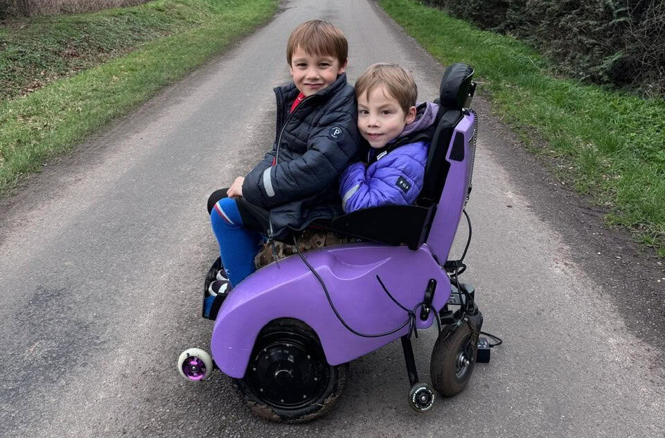 Otis sitting on Frankie's knees, on Frankie's purple electric wheelchair. They are both smiling widely.