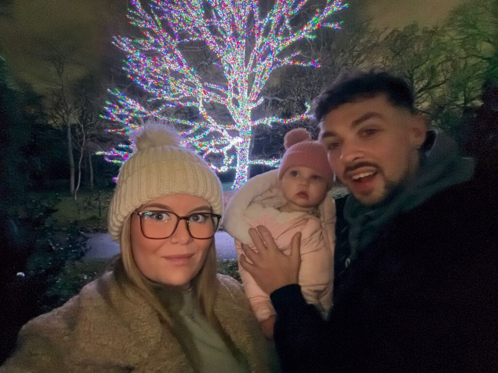Paige, her daughter, and her partner are smiling widely in front of a luminescent tree behind them.