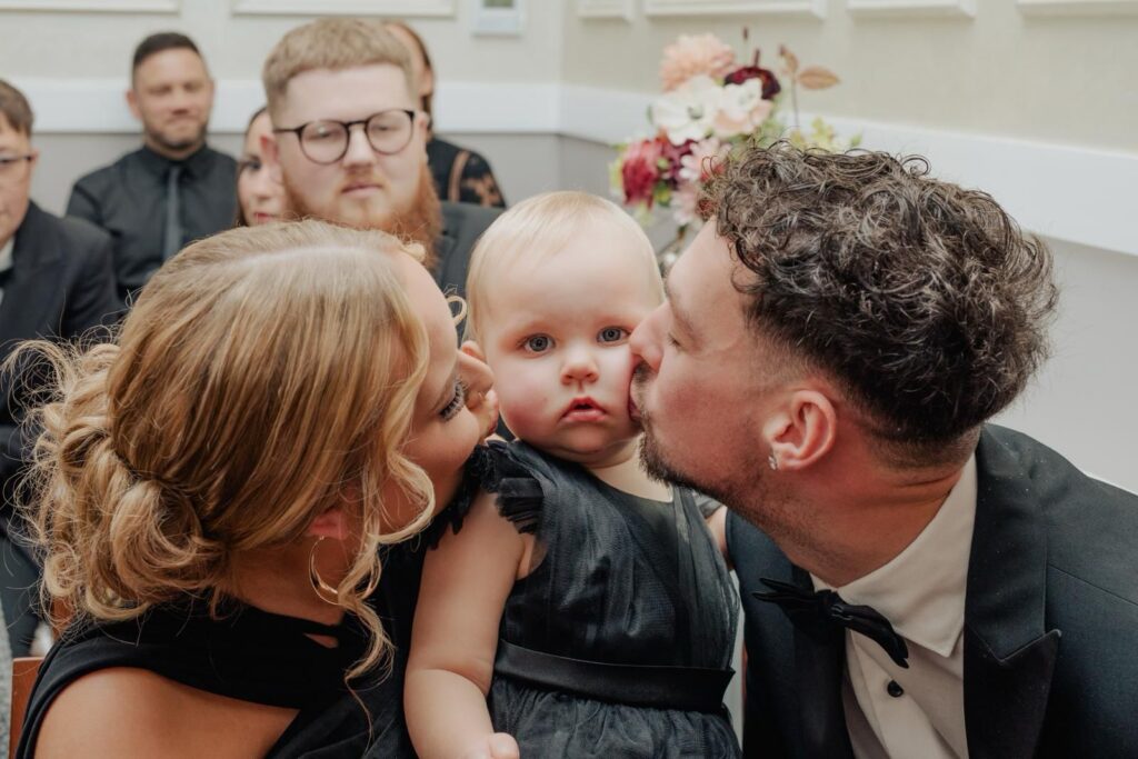 A mother and father lean in to kiss their daughter on either side of her cheeks. They are all wearing black coloured smart attire.